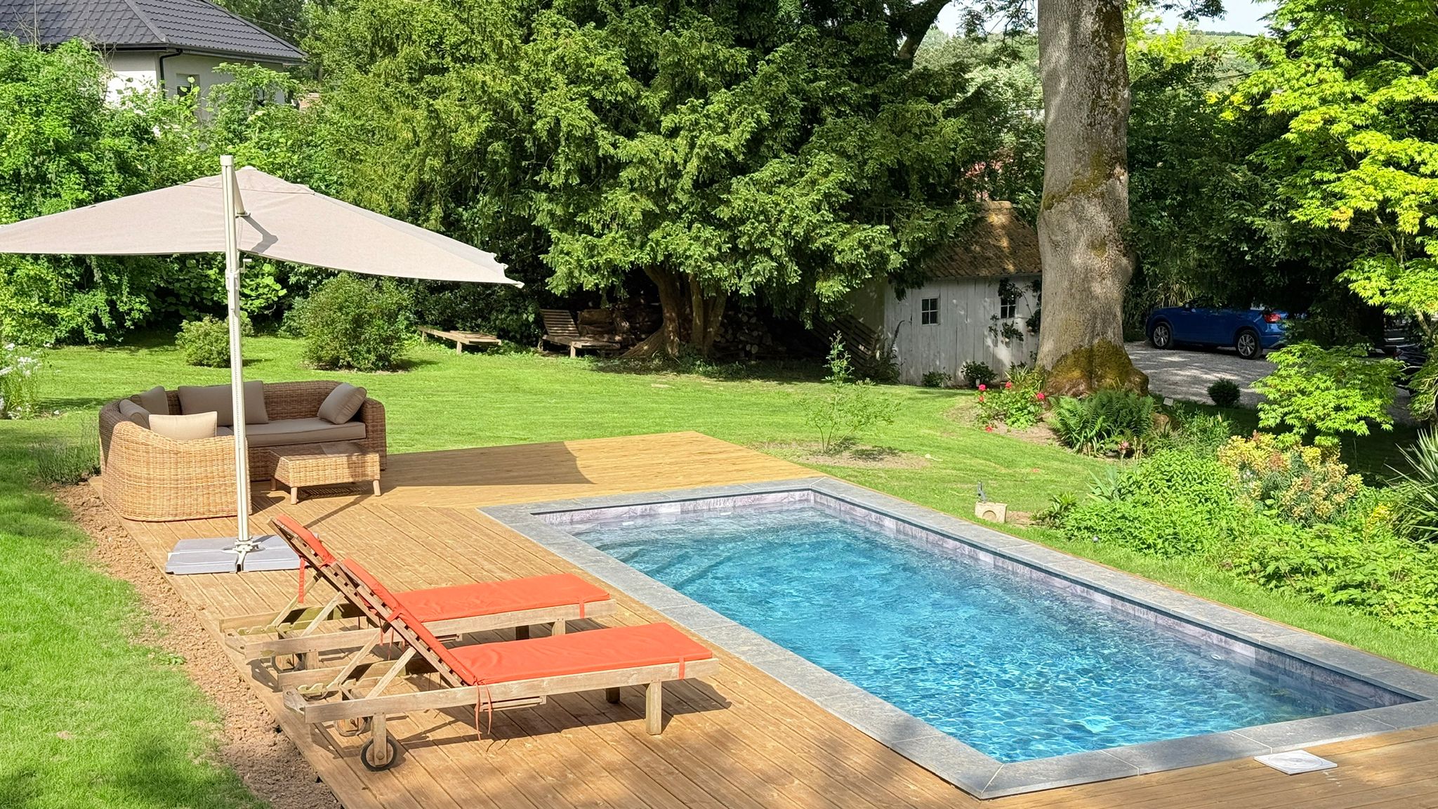 Une oasis de sérénité dans le jardin comprend une piscine rectangulaire, une terrasse en bois avec des chaises longues et un coin salon en osier sous un grand parasol. De l'herbe verte, des arbres et un petit bâtiment complètent cette scène idyllique.