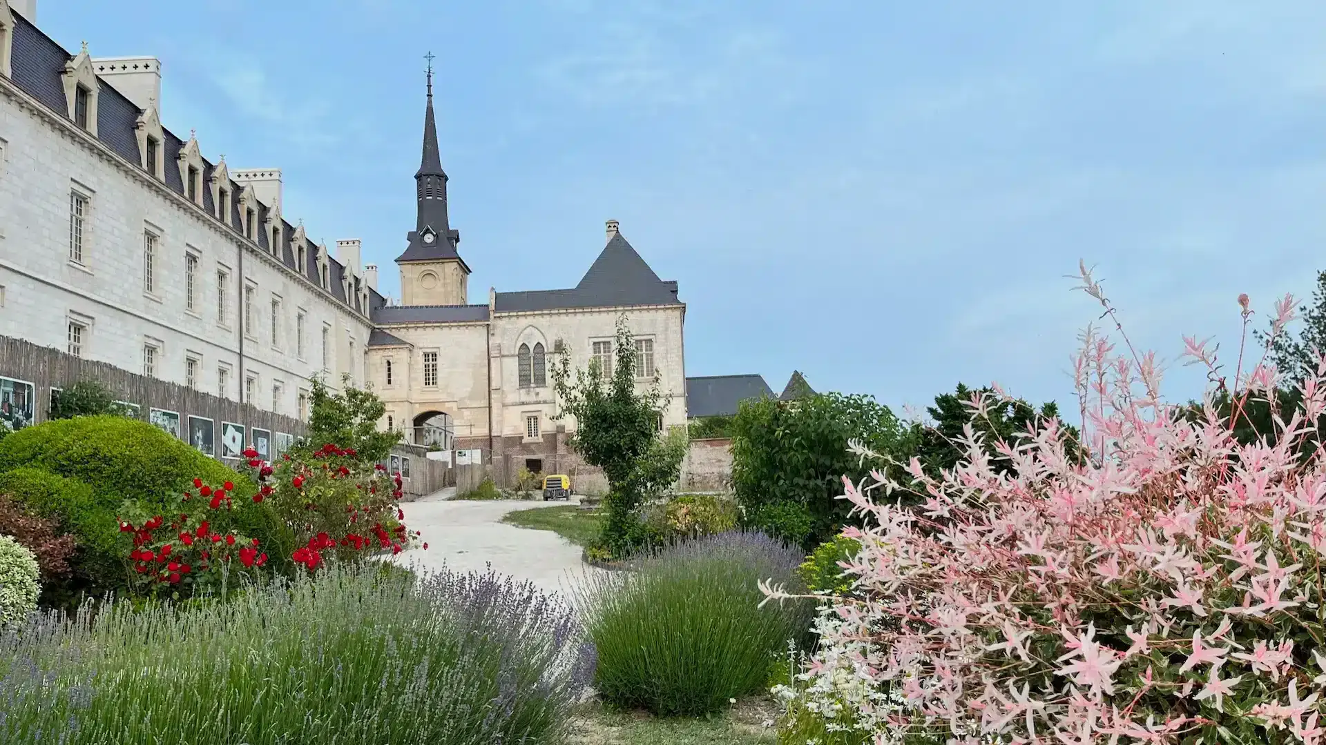 A stone chateau, La Plonplonière, with a tall spire stands beside a garden path lined with blooming flowers and purple lavender—a charming maison d’hôtes en Côte d’Opale under a clear blue sky.