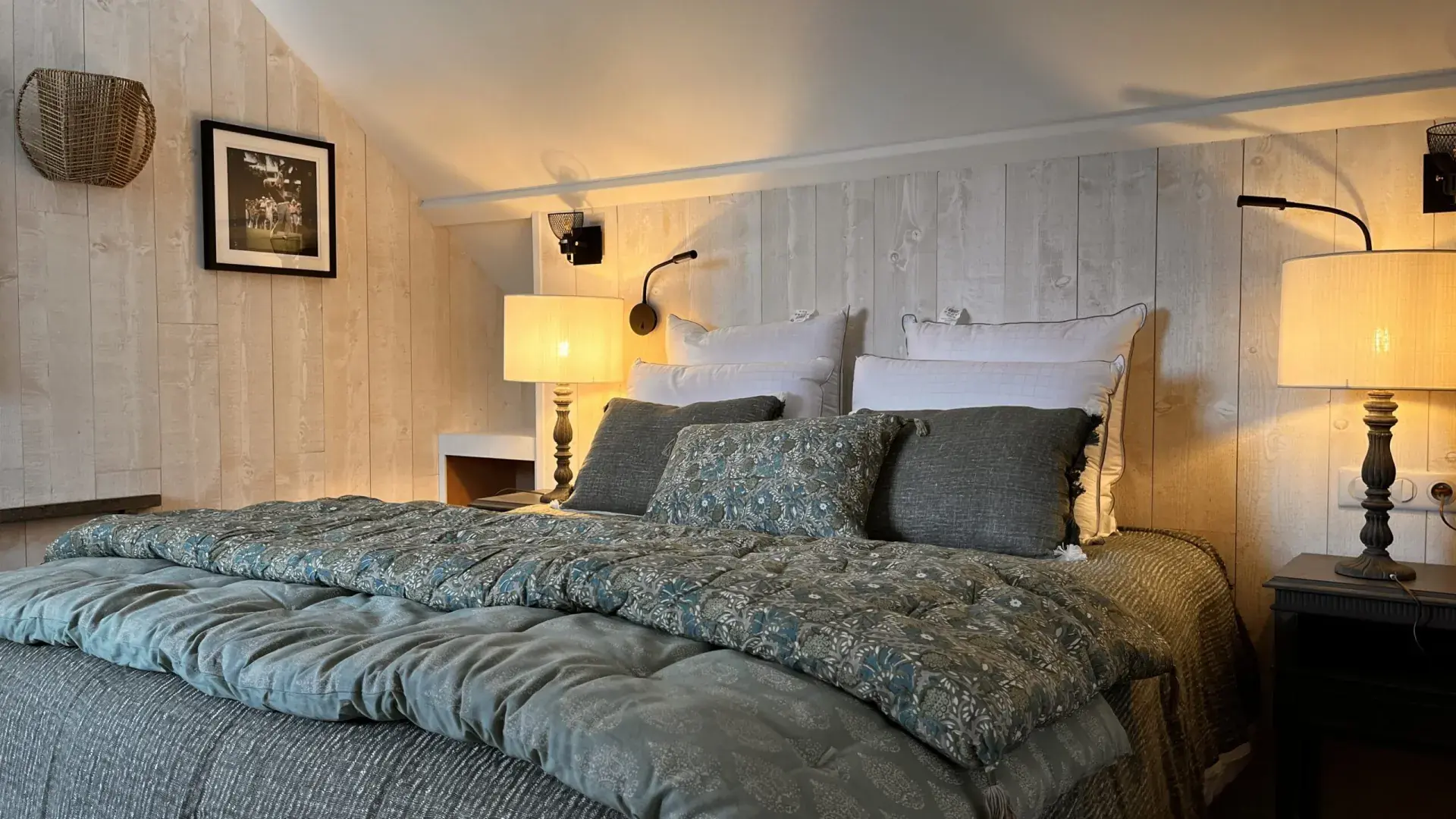 Cozy bedroom at La Plonplonière, a charming maison d'hôtes en Côte d'Opale, with a neatly made bed in green and gray, two pillows, patterned cushions, lamps on bedside tables, and a framed photo on the light wood-paneled wall.