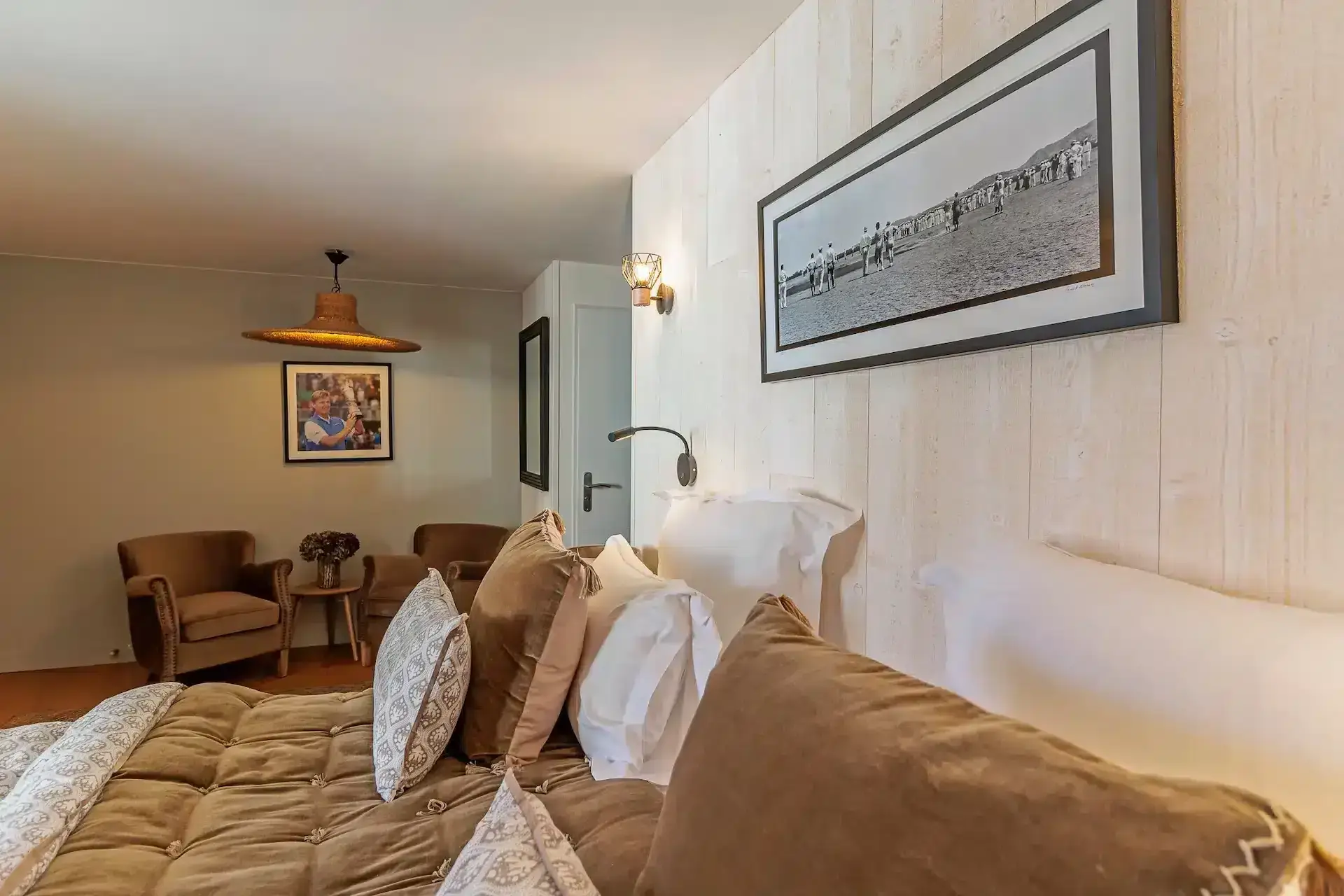 Cozy bedroom at La Plonplonière with a neatly made bed, brown and white pillows, two beige armchairs near a small round table, wall art, and soft ambient lighting—perfect for a warm stay in our Maison d'hôtes en Côte d'Opale.