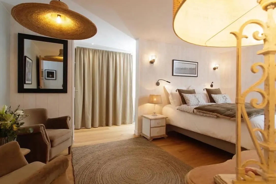 Cozy bedroom at La Plonplonière with a large bed, beige armchairs, round rug, and neutral curtains. Warm lighting creates an inviting atmosphere in this charming maison d’hôtes en Côte d’Opale.