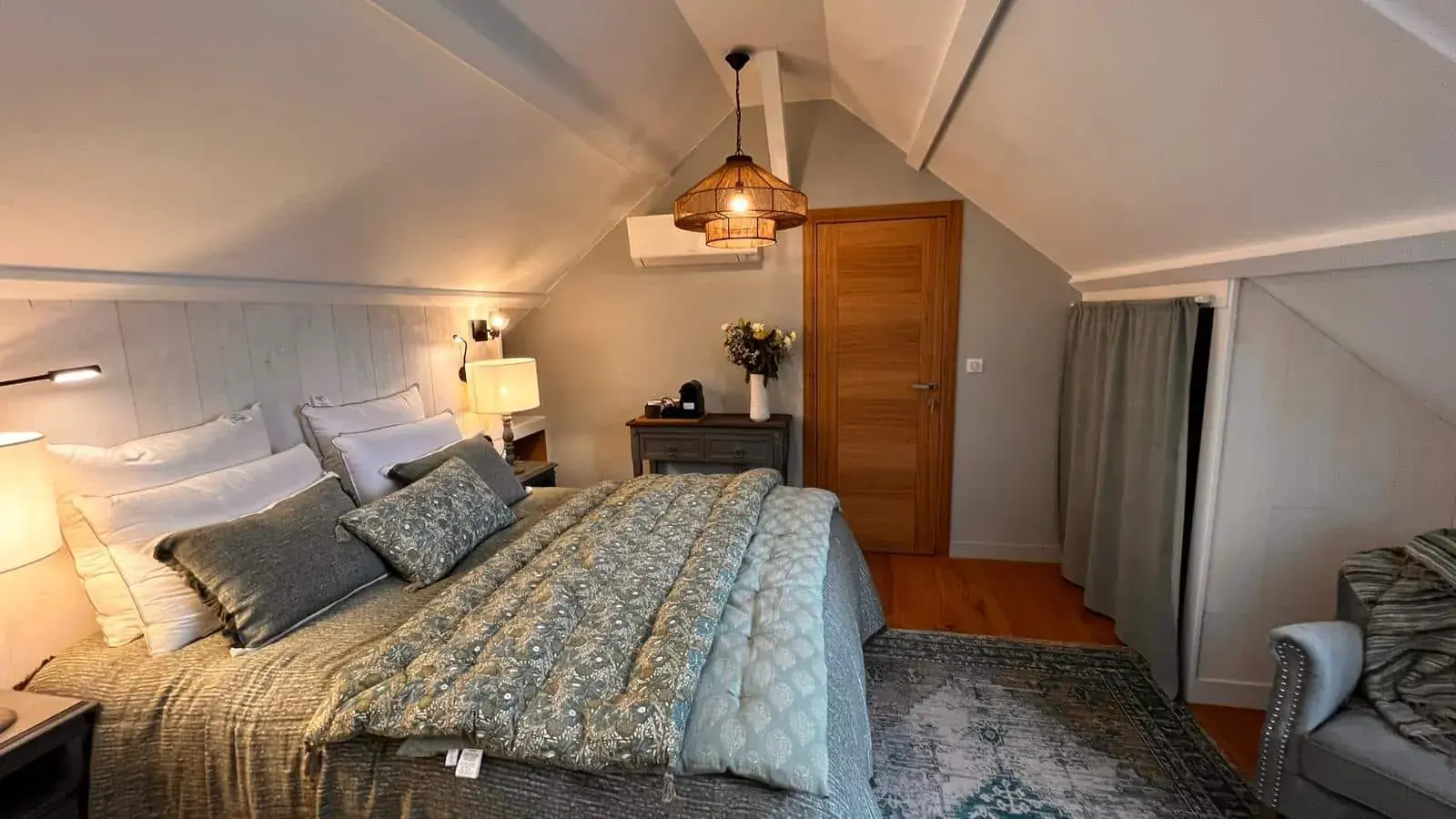 A cozy attic bedroom at La Plonplonière, maison d'hôtes en Côte d'Opale, with sloped ceilings, a large bed in blue and gray bedding, bedside lamps, dresser with flowers, wooden door, armchair on a rug, and warm relaxing light.
