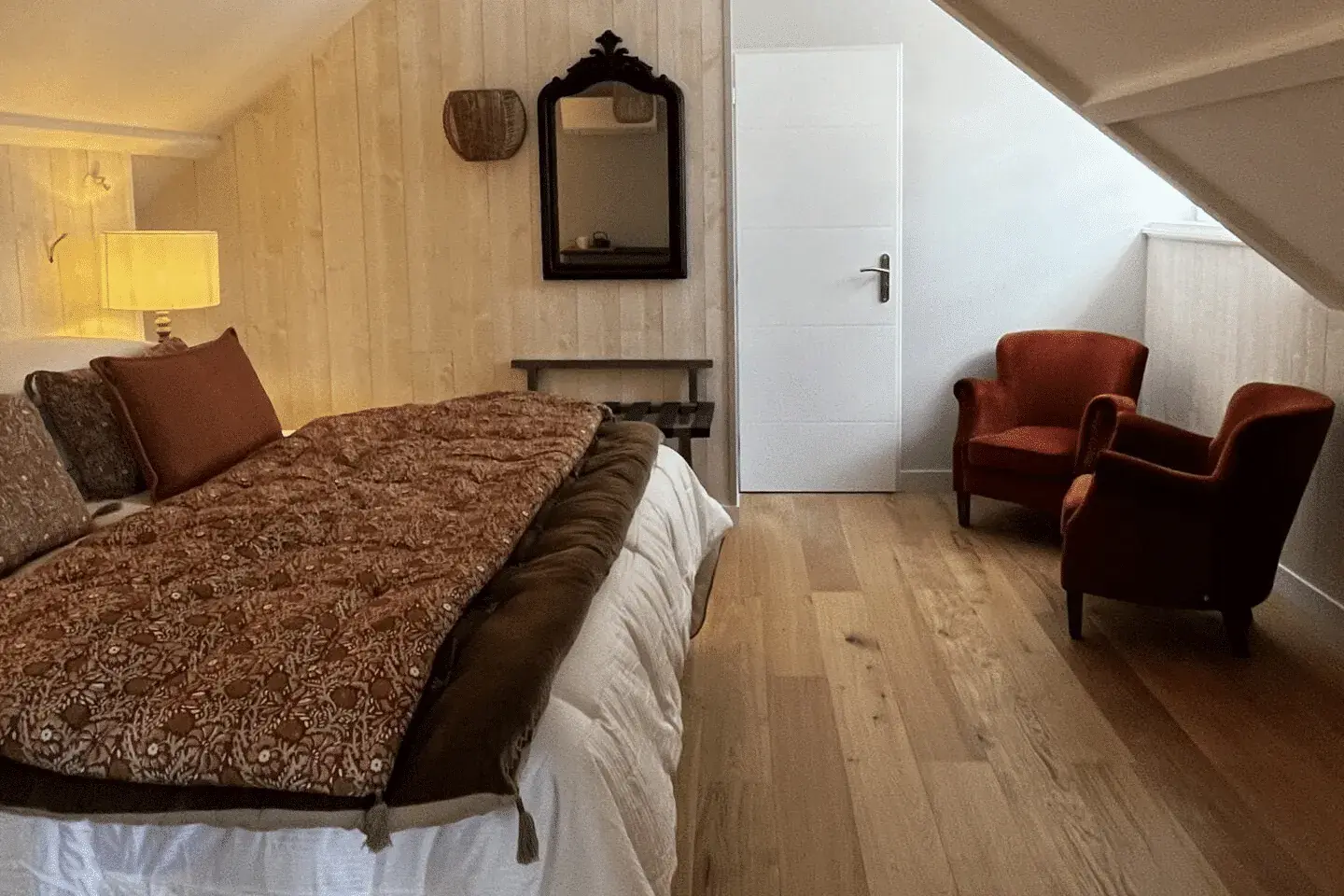 A cozy attic bedroom at La Plonplonière with a large bed, patterned blankets and pillows, wooden floor, wall mirror, small table, lamp, and two red armchairs by the slanted ceiling window—your ideal Maison d’hôtes en Côte d’Opale retreat.