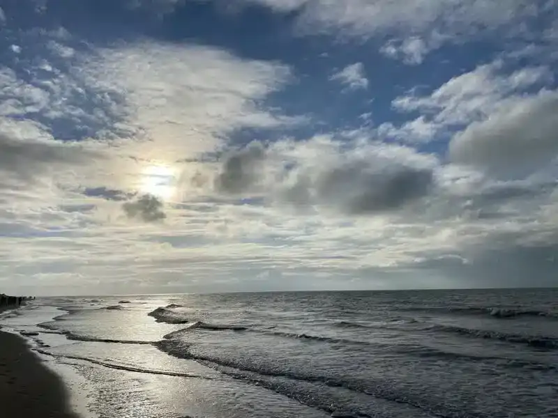 Un ciel nuageux avec un soleil partiellement visible brille sur une mer calme près de La Plonplonière. De douces vagues roulent sur une plage de sable et la lumière du soleil se reflète sur l'eau - une sérénité parfaite pour cette charmante maison d'hôtes en Côte d'Opale.