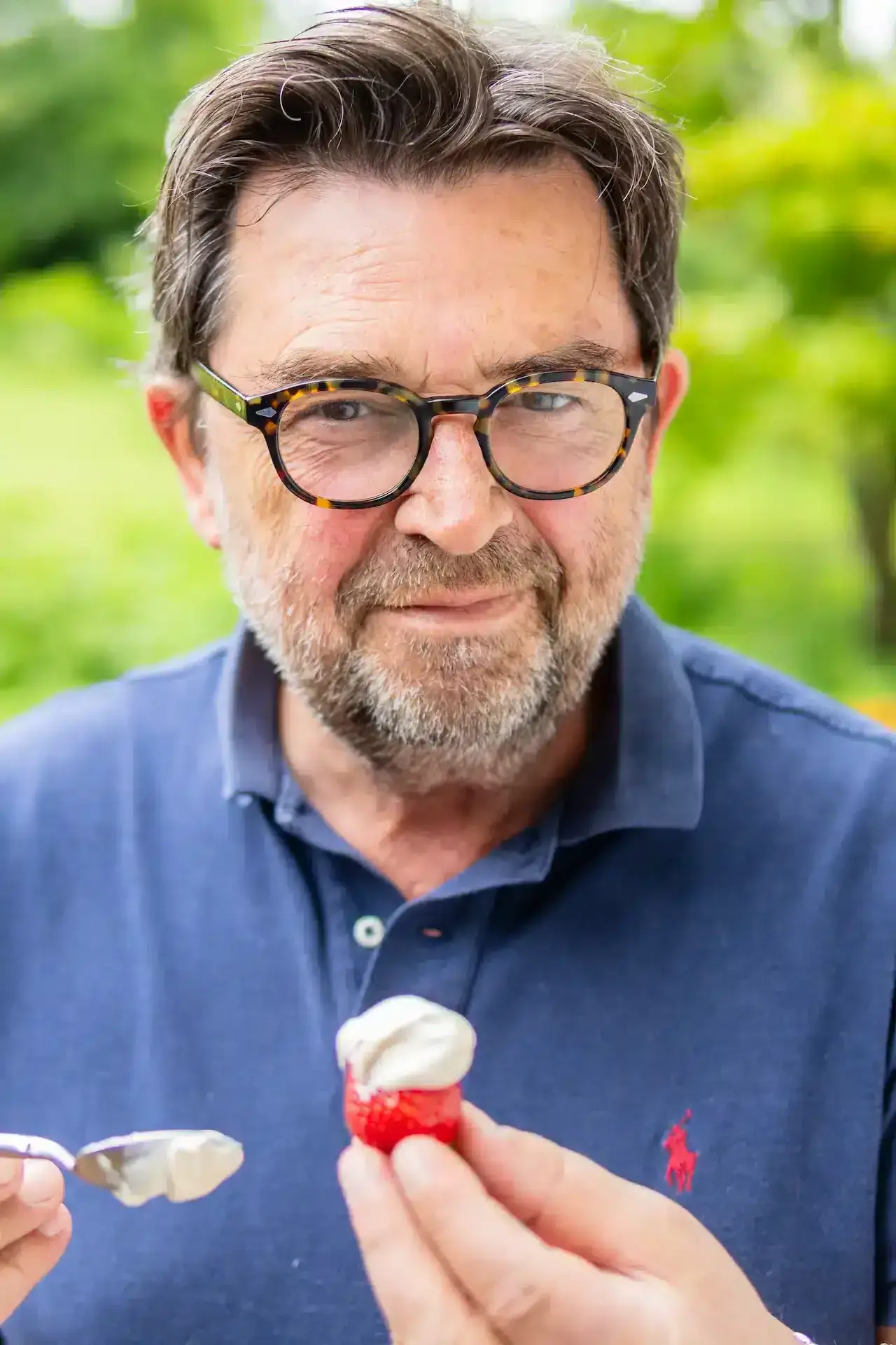 A middle-aged man with glasses and a beard stands outdoors at La Plonplonière, a charming maison d'hôtes en Côte d'Opale, smiling slightly as he holds a strawberry topped with cream and a spoon in each hand. He wears a blue polo shirt.
