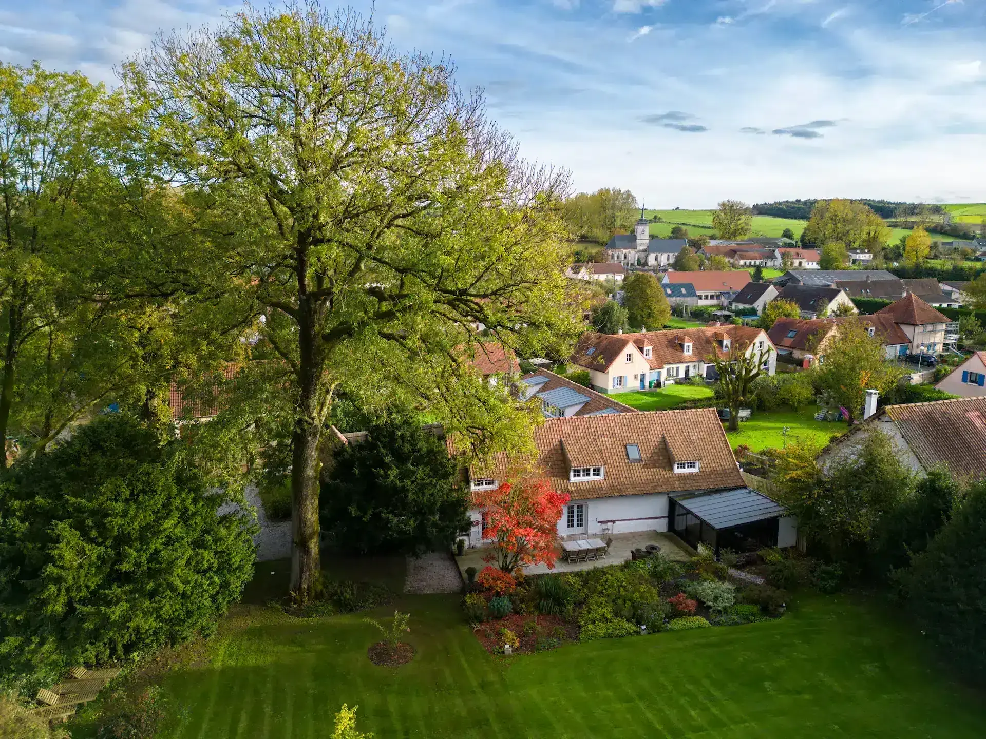Aerial view of a suburban neighborhood with houses, gardens, and large trees. In the foreground, La Plonplonière stands out—a Maison d’hôtes en Côte d’Opale with a beige roof, glass-covered patio, and lush well-kept lawn.