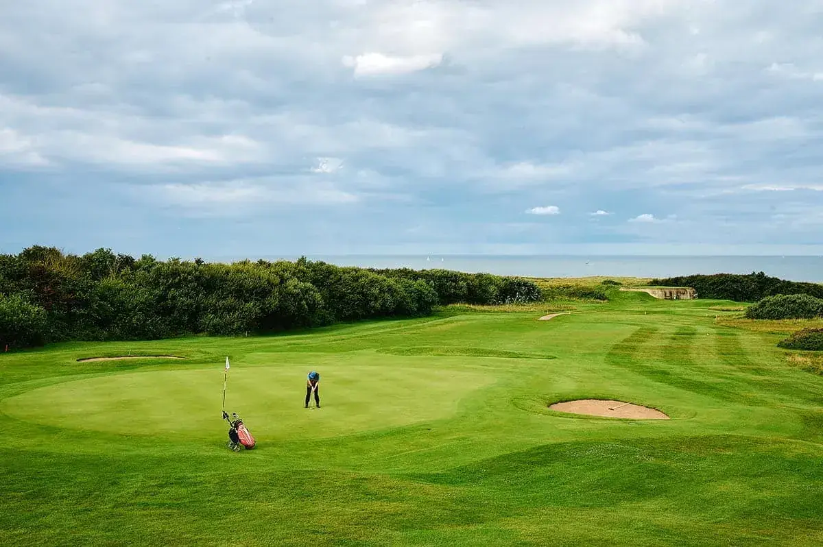 A golfer stands on a green golf course near the hole at La Plonplonière, a Maison d'hôtes en Côte d'Opale, surrounded by lush grass, sand bunkers, and trees, with a cloudy sky and distant sea in the background.