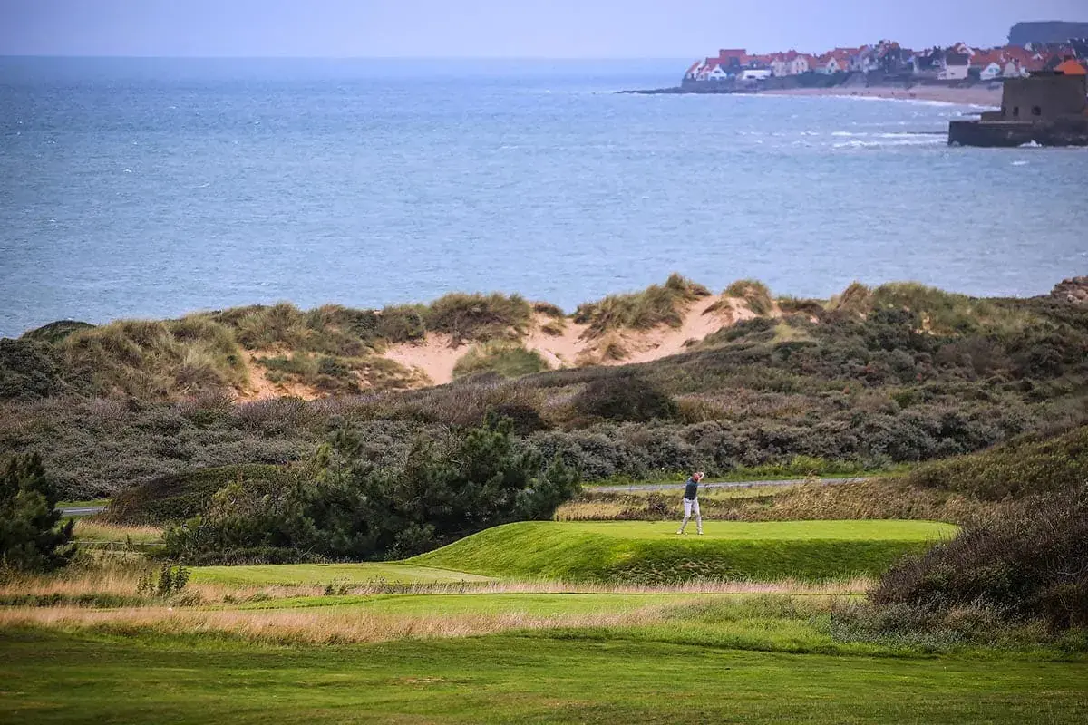 A golfer stands on a green fairway near grassy dunes, with the sea and a distant coastal village in view—perfect scenery for guests staying at La Plonplonière, charmante maison d’hôtes en Côte d’Opale under a cloudy sky.