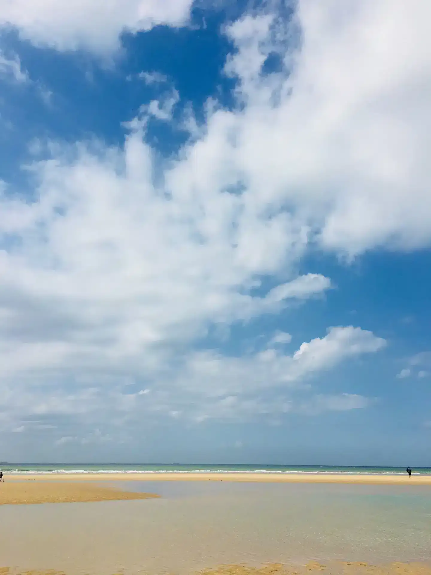 A peaceful beach scene near La Plonplonière, with shallow water, sandy shore, and a vast sky filled with fluffy white clouds. The calm ocean and distant horizon evoke the charm of chambres d'hôtes en Côte d'Opale.