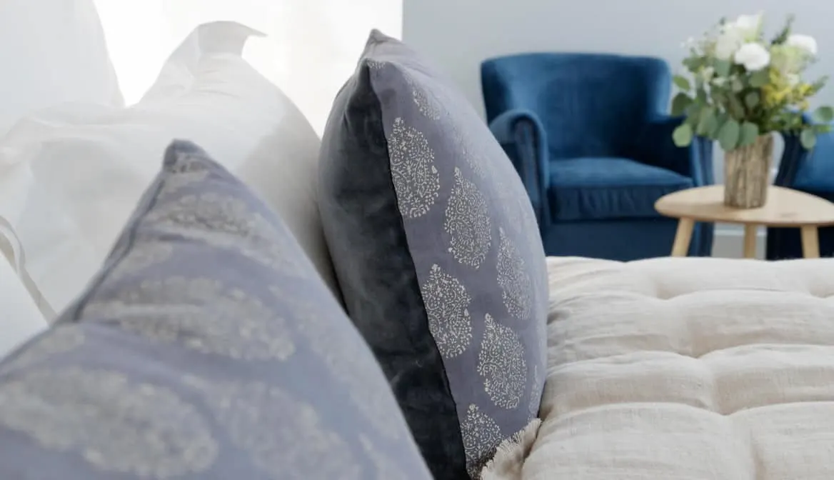 Close-up of a bed with white sheets and two decorative gray pillows with patterns at La Plonplonière, une charmante maison d’hôtes en Côte d’Opale. In the background, a blue armchair and a small round table with a vase of white flowers are visible.