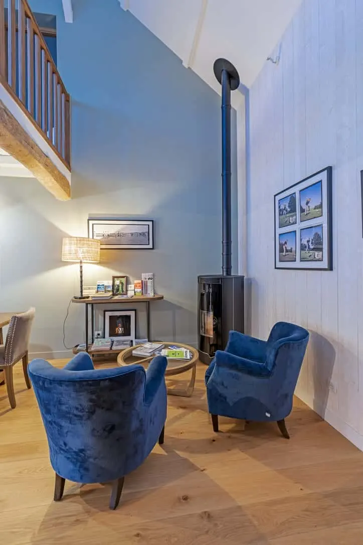 Le salon confortable de La Plonplonière comprend deux fauteuils en velours bleu autour d'une petite table, un poêle à bois noir, des tableaux encadrés et un escalier - parfait pour les clients qui recherchent des chambres d'hôtes en Côte d'Opale.