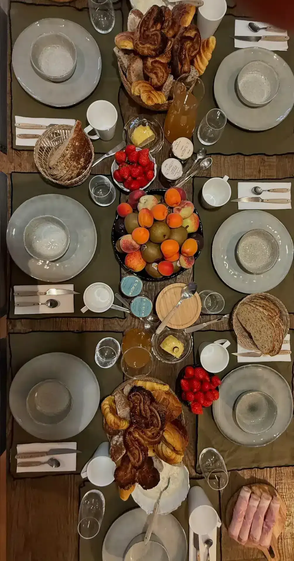 A breakfast table at La Plonplonière, maison d'hôtes en Côte d'Opale, set for six with plates, cutlery, fresh bread, croissants, fruits, butter, jams, orange juice, and coffee cups—all beautifully arranged and viewed from above.