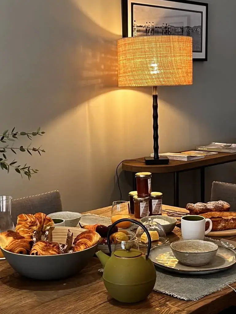 A cozy breakfast setting at La Plonplonière, a charming maison d'hôtes en Côte d'Opale, with a green teapot, mugs, fresh pastries, jams, and a warm lamp-lit ambiance for a perfect start to your day.
