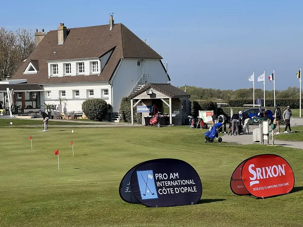 A golf course with a clubhouse in the background, people golfing, and banners for Pro Am International Côte d’Opale and Srixon. White flags fly near the building—perfect for guests at La Plonplonière, a charming maison d'hôtes en Côte d'Opale.