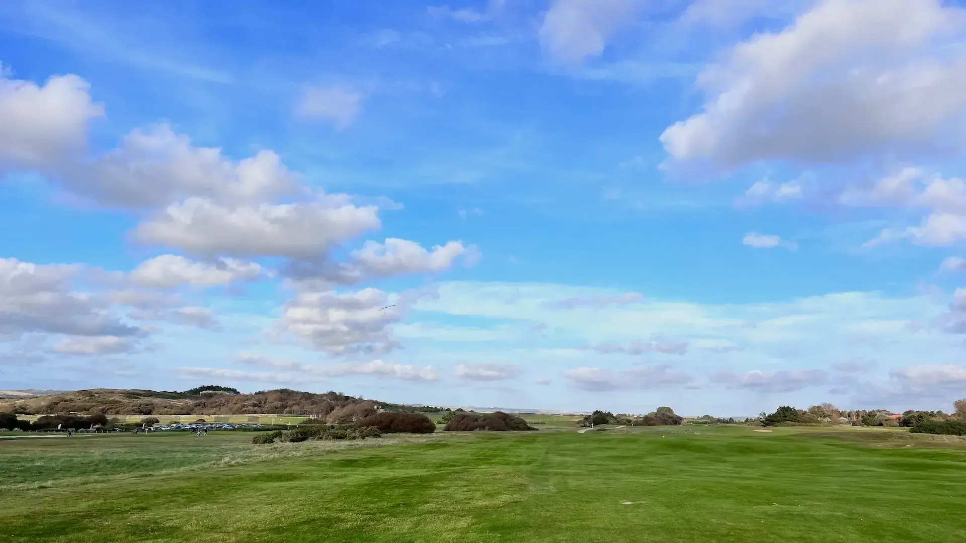 A wide, open golf course with bright green grass under a blue sky dotted with fluffy clouds, just beyond the peaceful Chambres d'hôtes en Côte d'Opale at La Plonplonière. Low hills and scattered bushes grace the distance.
