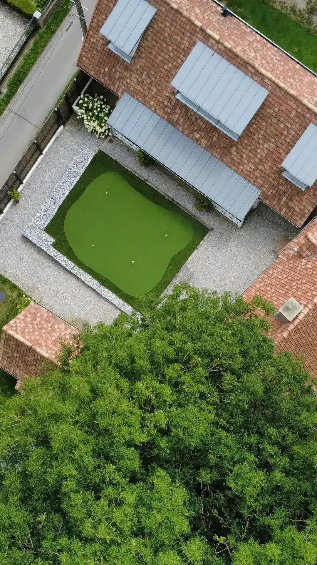 Vue aérienne de La Plonplonière, une Maison d'hôtes en Côte d'Opale, avec un petit putting green entouré de gravier dans une cour entre des bâtiments aux tuiles rouges, à côté d'un arbre feuillu.