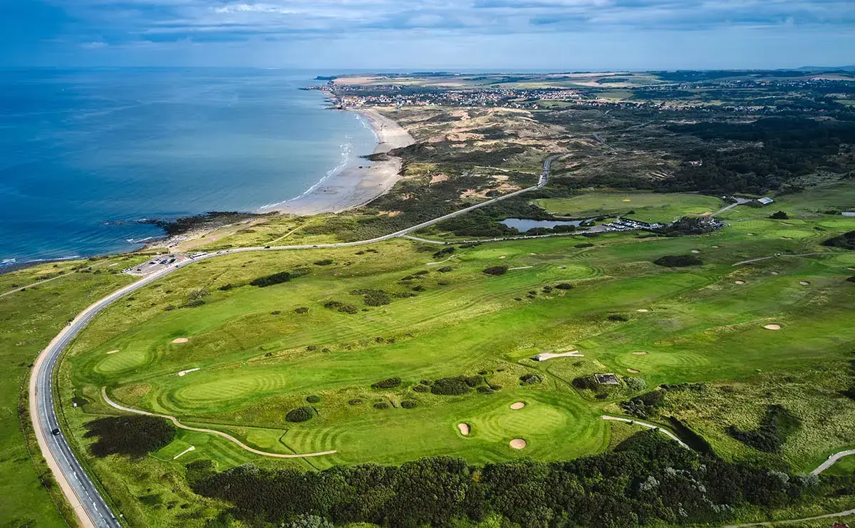Aerial view of a coastal golf course near La Plonplonière, with green fairways, sand bunkers, and a winding road. The sea and sandy beach border the Maison d'hôtes en Côte d'Opale under a partly cloudy sky.