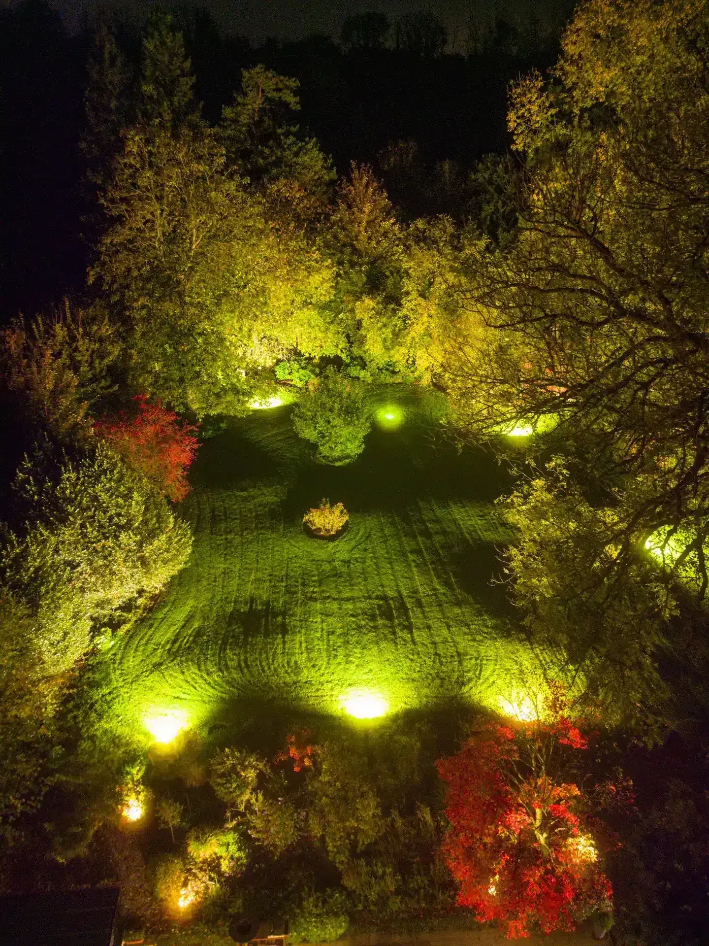 An aerial view of La Plonplonière, a Maison d’hôtes en Côte d’Opale, shows its garden at night: spotlights cast a warm yellow glow on grass and trees, while red-tinged leaves add vibrant color to this inviting Chambres d’hôtes en Côte d’Opale scene.